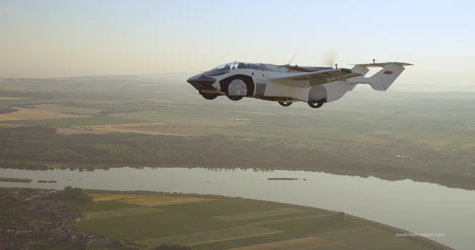White and black AirCar flying car with wings extended, flying over farmland and a river